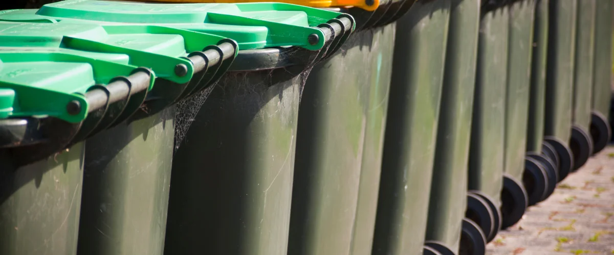 A close-up, linear view of several green wheelie bins with bright green lids, showing some cobwebs on the side. A close-up, linear view of several green wheelie bins with bright green lids, showing some cobwebs on the side.