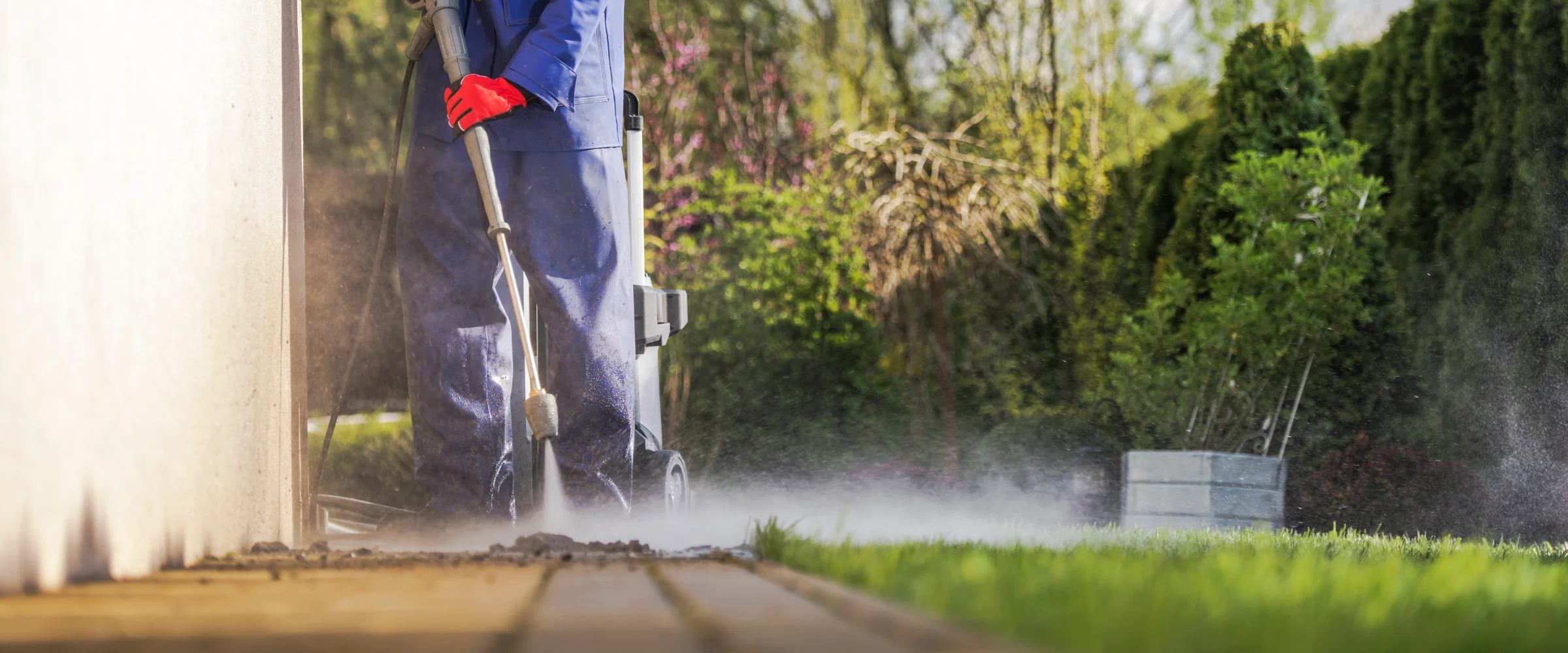 A person in blue coveralls and red gloves pressure washing a dirty outdoor patio, with water spray and green plants. A person in blue coveralls and red gloves pressure washing a dirty outdoor patio, with water spray and green plants.
