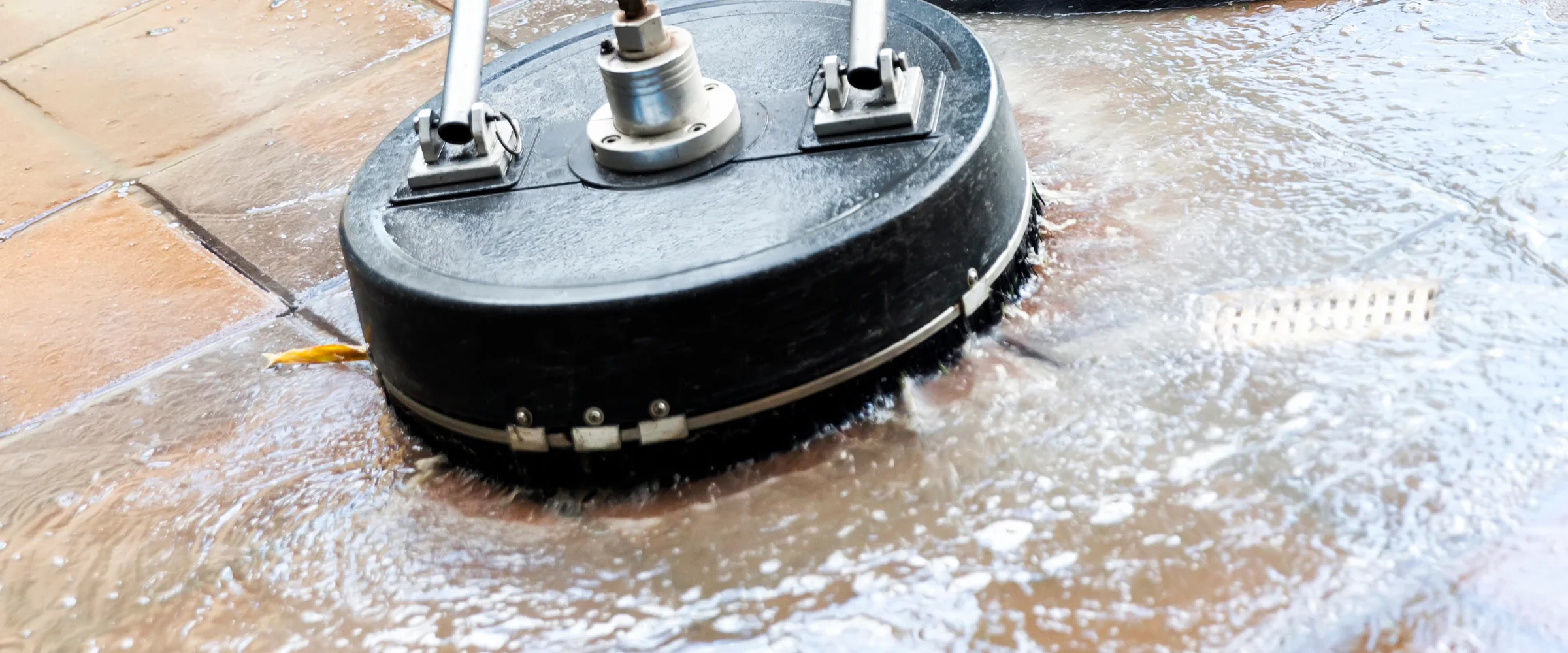A close-up of a black circular surface cleaner attachment actively washing a tiled patio, splashing water and dirt. A close-up of a black circular surface cleaner attachment actively washing a tiled patio, splashing water and dirt.