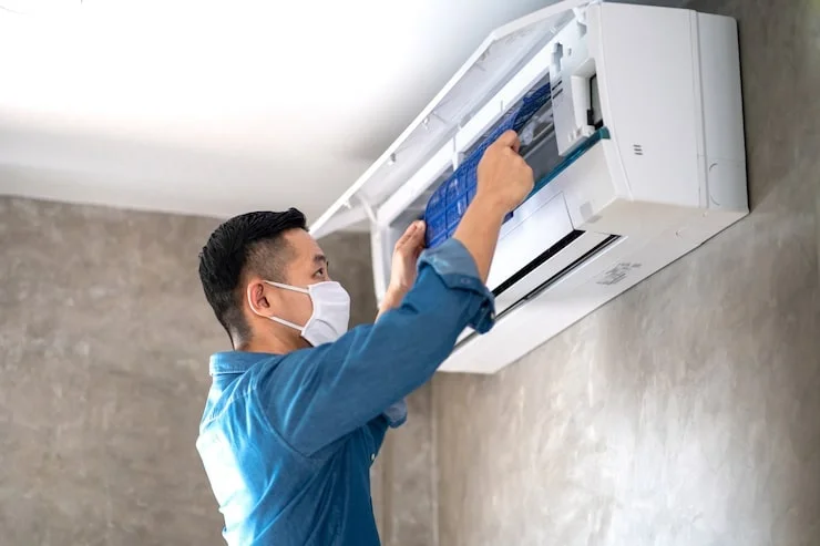 A masked man in a denim shirt adjusts a blue filter inside a wall-mounted air conditioner unit.