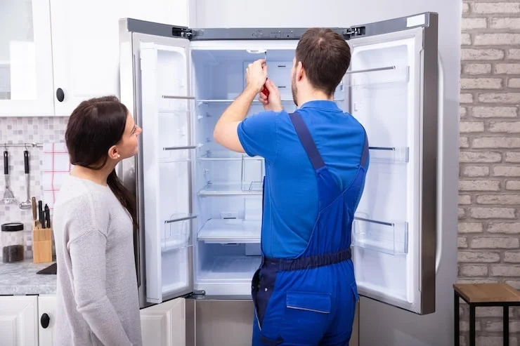 A woman watches as a service technician in blue overalls repairs the inside of a stainless steel refrigerator.