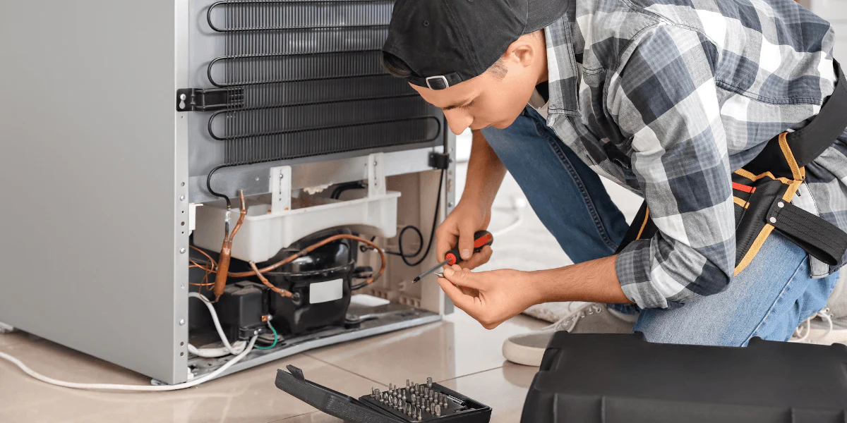 A bearded man in a black cap and safety glasses, wearing gloves, cleans the filters of an air conditioning unit.