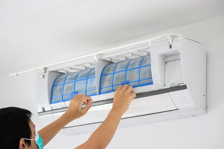 A person wearing a face mask cleans dirty air conditioner filters in a white wall-mounted unit.