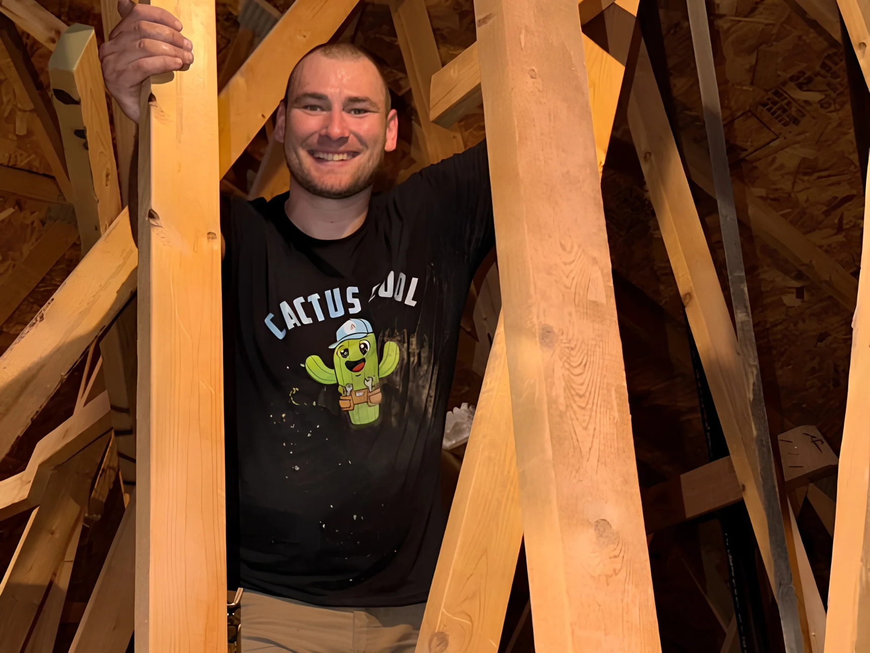 A smiling young man in a black 'Cactus Cool' shirt emerges from attic rafters, looking happy.