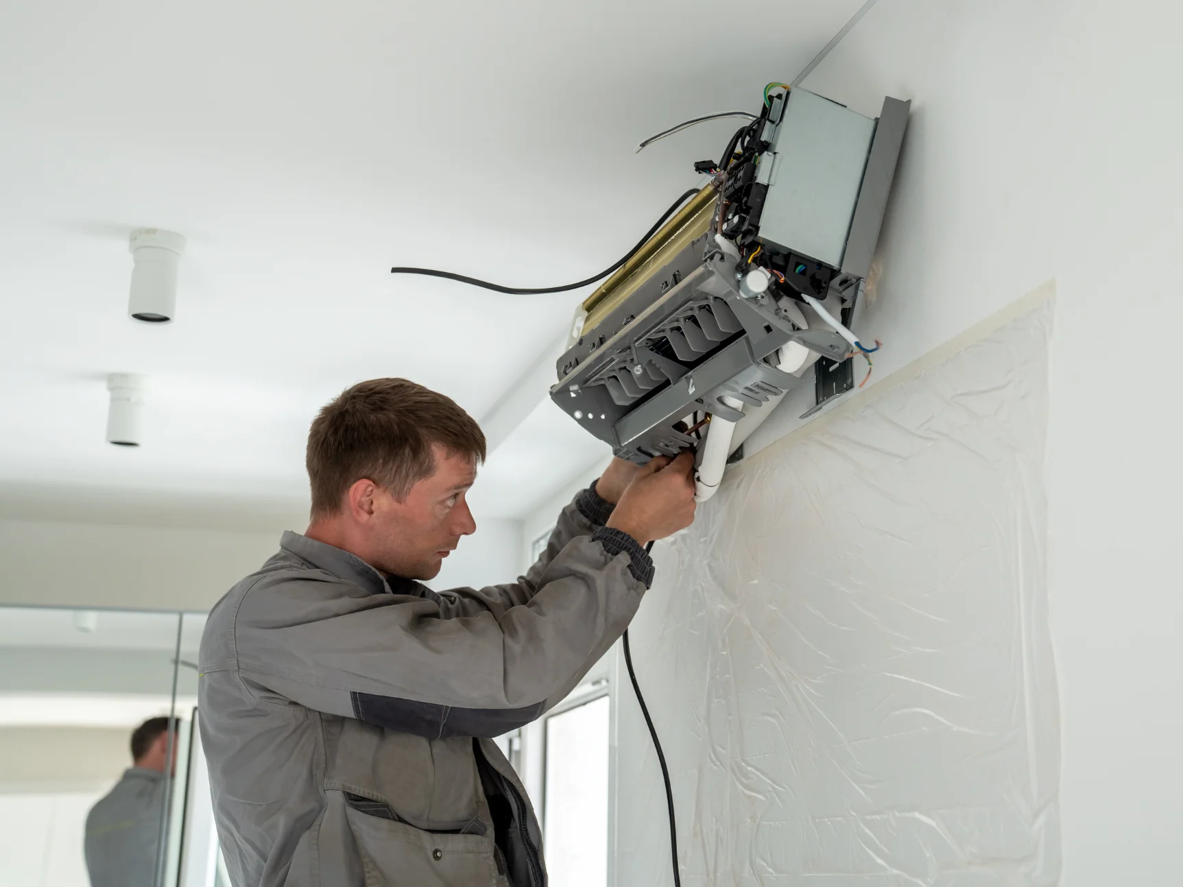 A professional HVAC repairman in a gray uniform installs an air conditioning unit on a white wall.