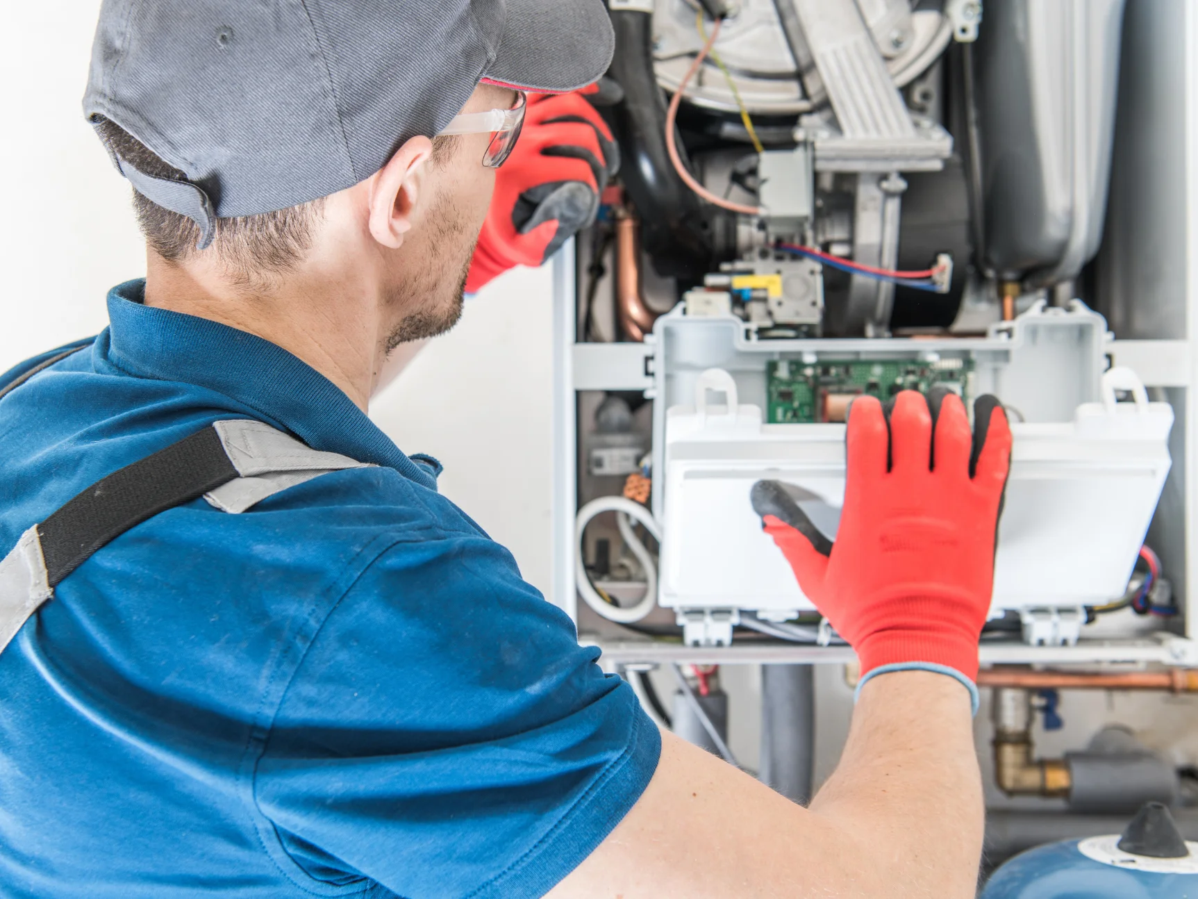 A HVAC technician in a blue shirt, grey cap, and safety glasses wearing red and black gloves works on a boiler unit.
