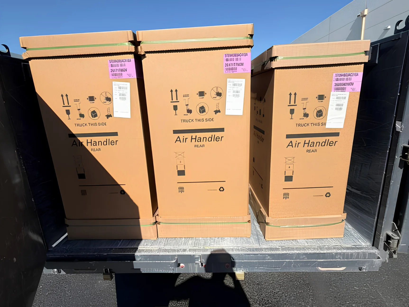 Three large cardboard boxes labeled 'Air Handler' strapped in a truck bed under a bright sky.
