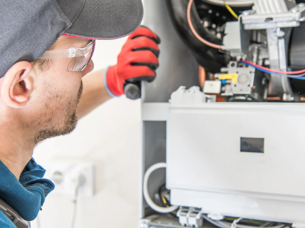 A technician in a cap and safety glasses, wearing a red glove, performing maintenance on a residential boiler.
