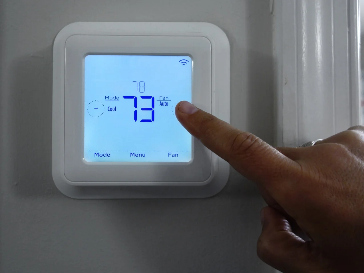 A hand pushing the auto fan button on a white digital thermostat displaying a 73-degree setting.