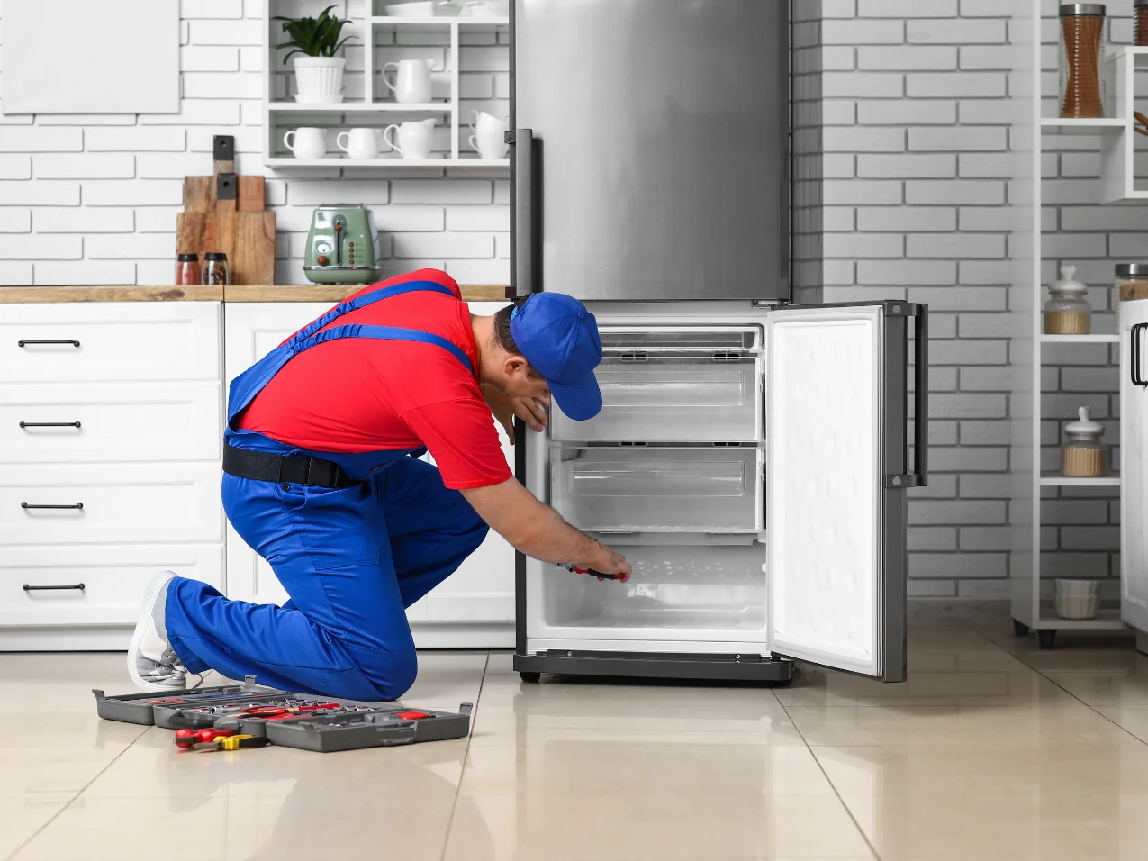 A service technician in blue overalls and a red shirt kneels and works on an open refrigerator in a modern kitchen.