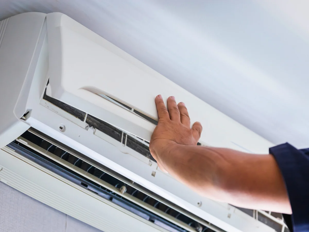 A close-up of a person's hand opening the cover of a dirty white air conditioning unit for maintenance.