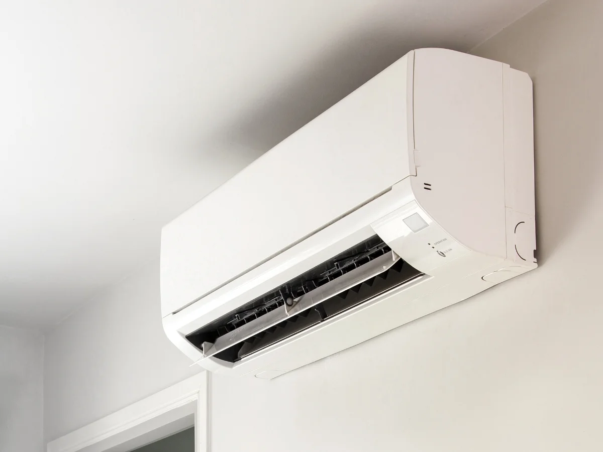 A white ductless mini-split air conditioner unit is mounted high on a light gray wall, seen from a low angle.