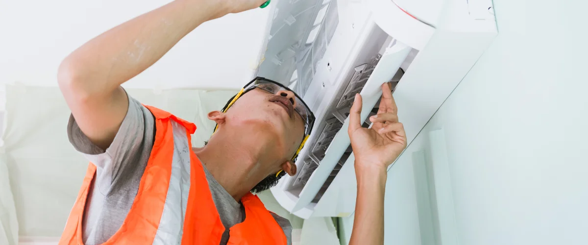 A technician in safety gear and orange vest performing maintenance on an air conditioner unit, viewed from below.