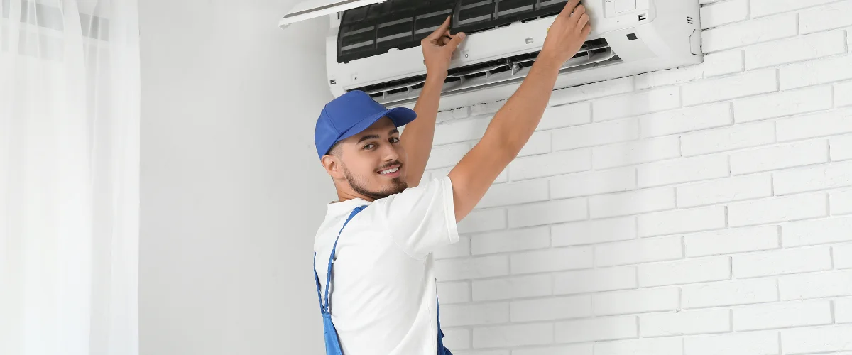A smiling male technician in a blue cap installing or repairing a wall-mounted air conditioner unit. A smiling male technician in a blue cap installing or repairing a wall-mounted air conditioner unit.
