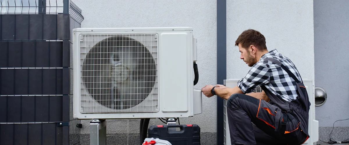 A male technician in work overalls inspecting an outdoor HVAC unit against a white wall. A male technician in work overalls inspecting an outdoor HVAC unit against a white wall.
