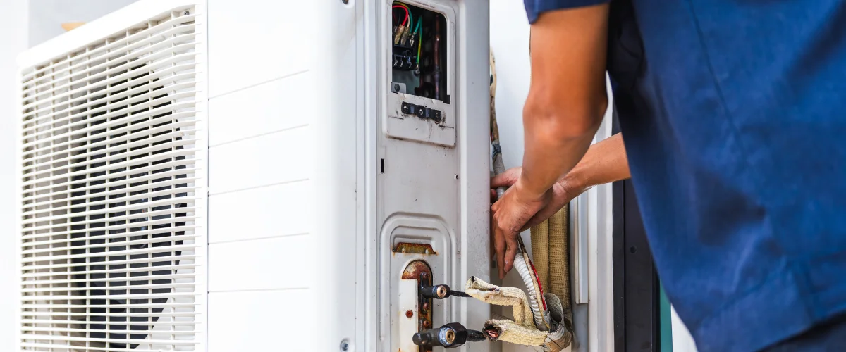A technician in a blue shirt adjusting wires and pipes on an open outdoor HVAC unit.