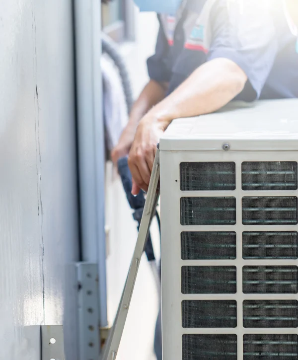 A service technician in uniform works on an outdoor air conditioning unit, with a focus on its grates and coils.
