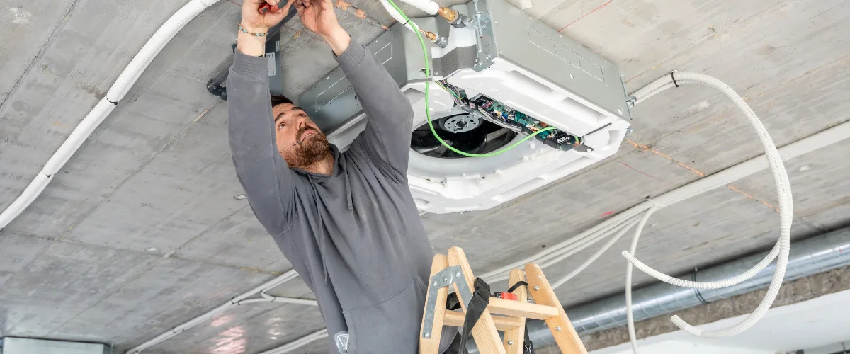 A bearded man in a gray hoodie on a ladder installs an HVAC unit into a concrete ceiling, surrounded by white conduits.