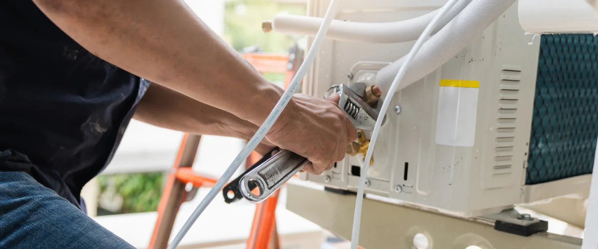 A close-up of a technician's hands using wrenches to install piping on an HVAC outdoor unit.