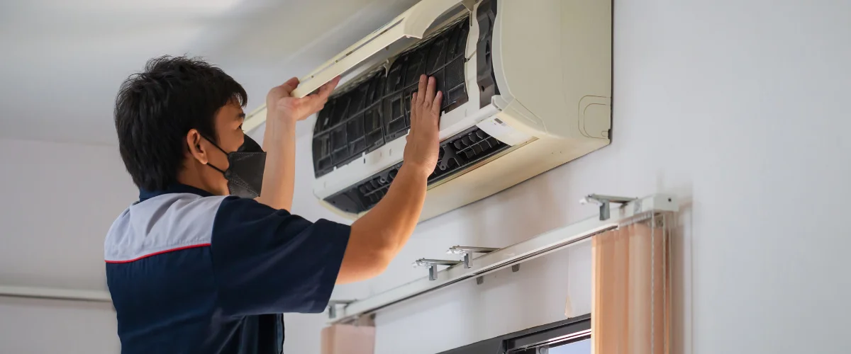 A technician from behind, wearing a navy shirt, connects copper pipes for an air conditioning unit with insulated lines.
