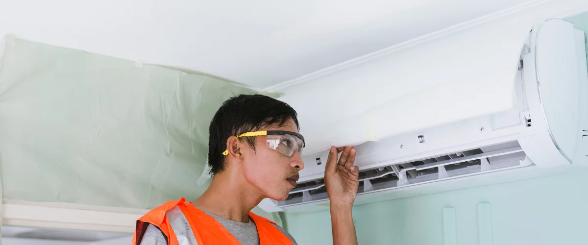 A technician wearing safety goggles and an orange vest inspects an open air conditioning unit on a wall.