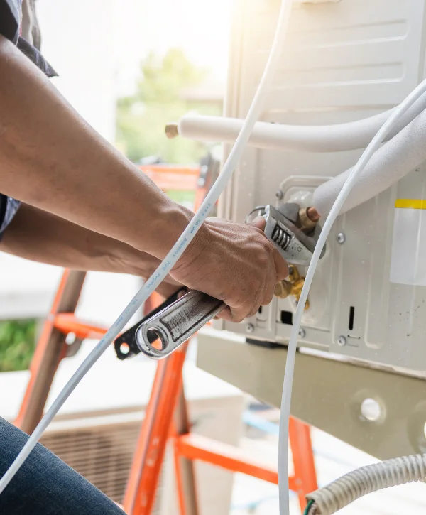 A technician from behind, wearing a navy shirt, connects copper pipes for an air conditioning unit with insulated lines.