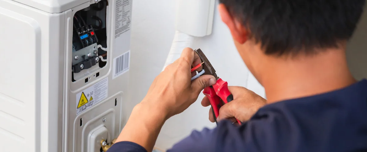 A technician from behind using red-handled pliers to strip wires on an outdoor air conditioning unit.