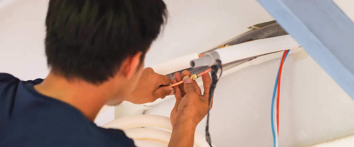 A technician from behind using red-handled pliers to strip wires on an outdoor air conditioning unit.