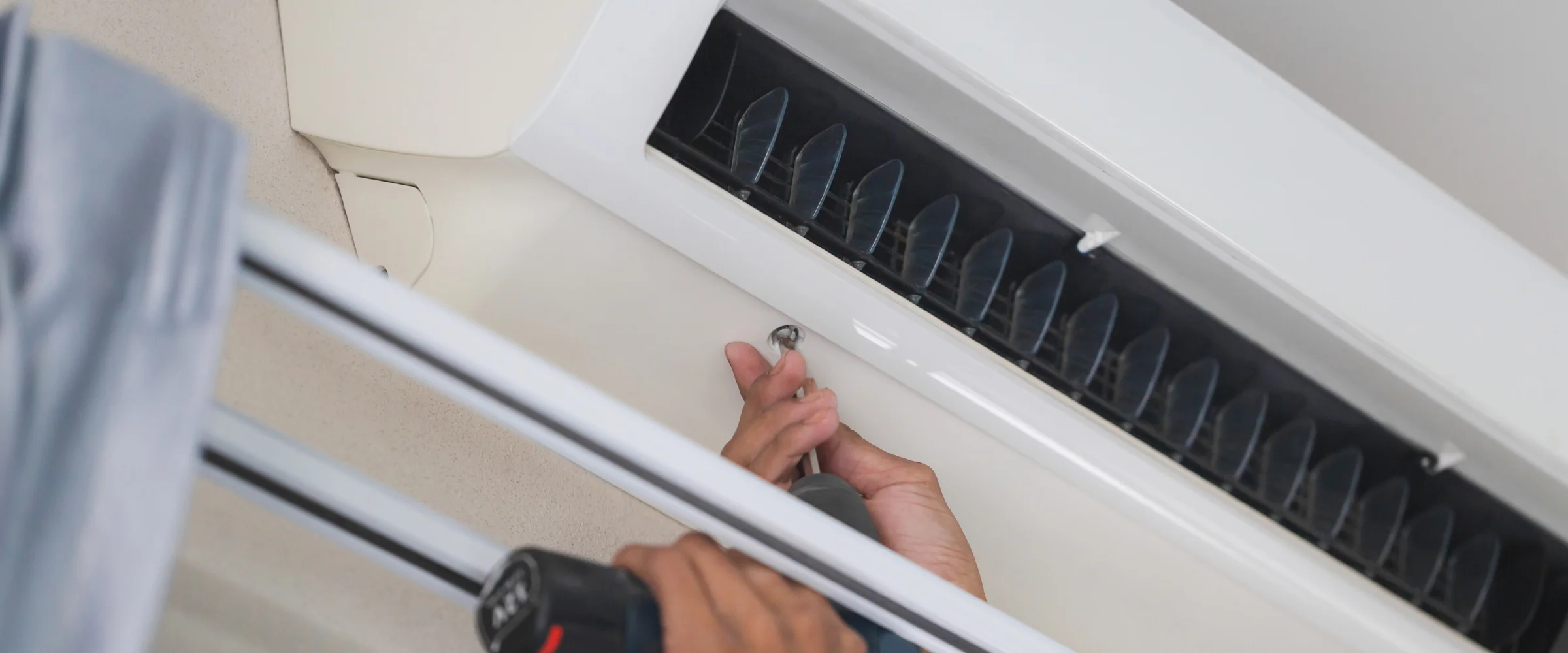 A gloved hand uses a screwdriver to perform maintenance on an air conditioner unit with visible filters.