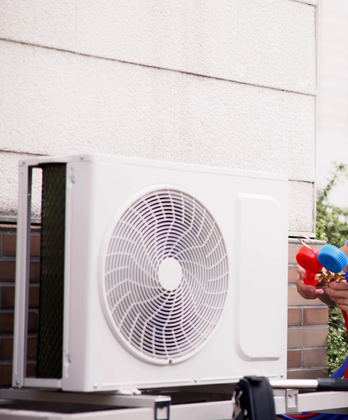 A white outdoor air conditioning unit with a person's hands holding red and blue gauges nearby, against a house wall.