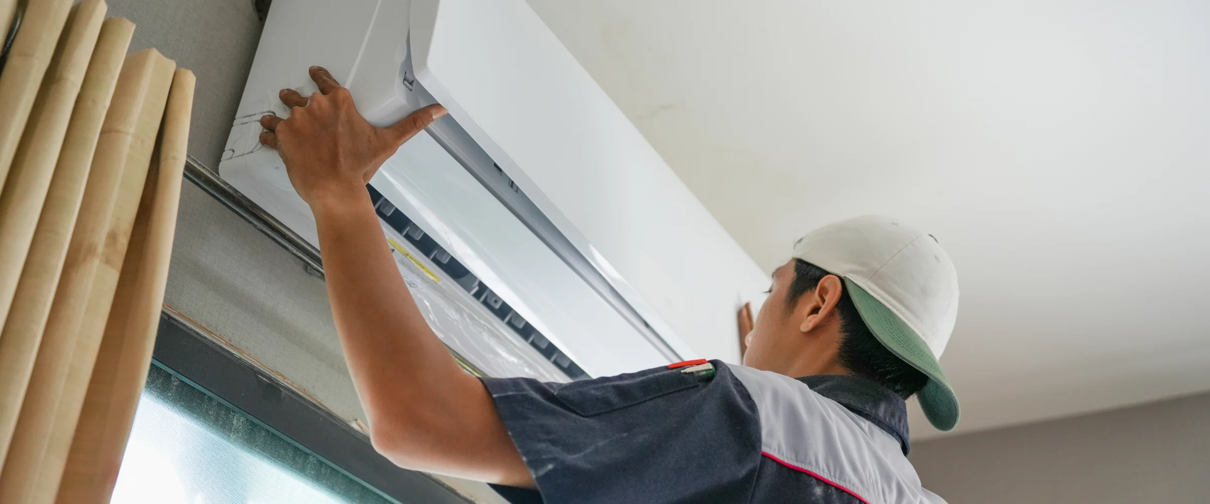 A technician in a baseball cap installing or repairing a white split-system air conditioner unit indoors.