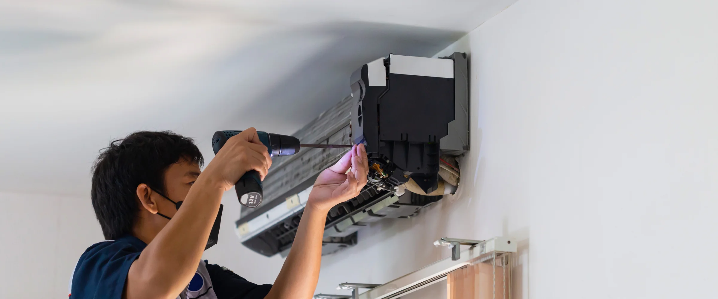 A technician wearing a black mask uses a power drill to repair or install an air conditioning unit on a white wall.