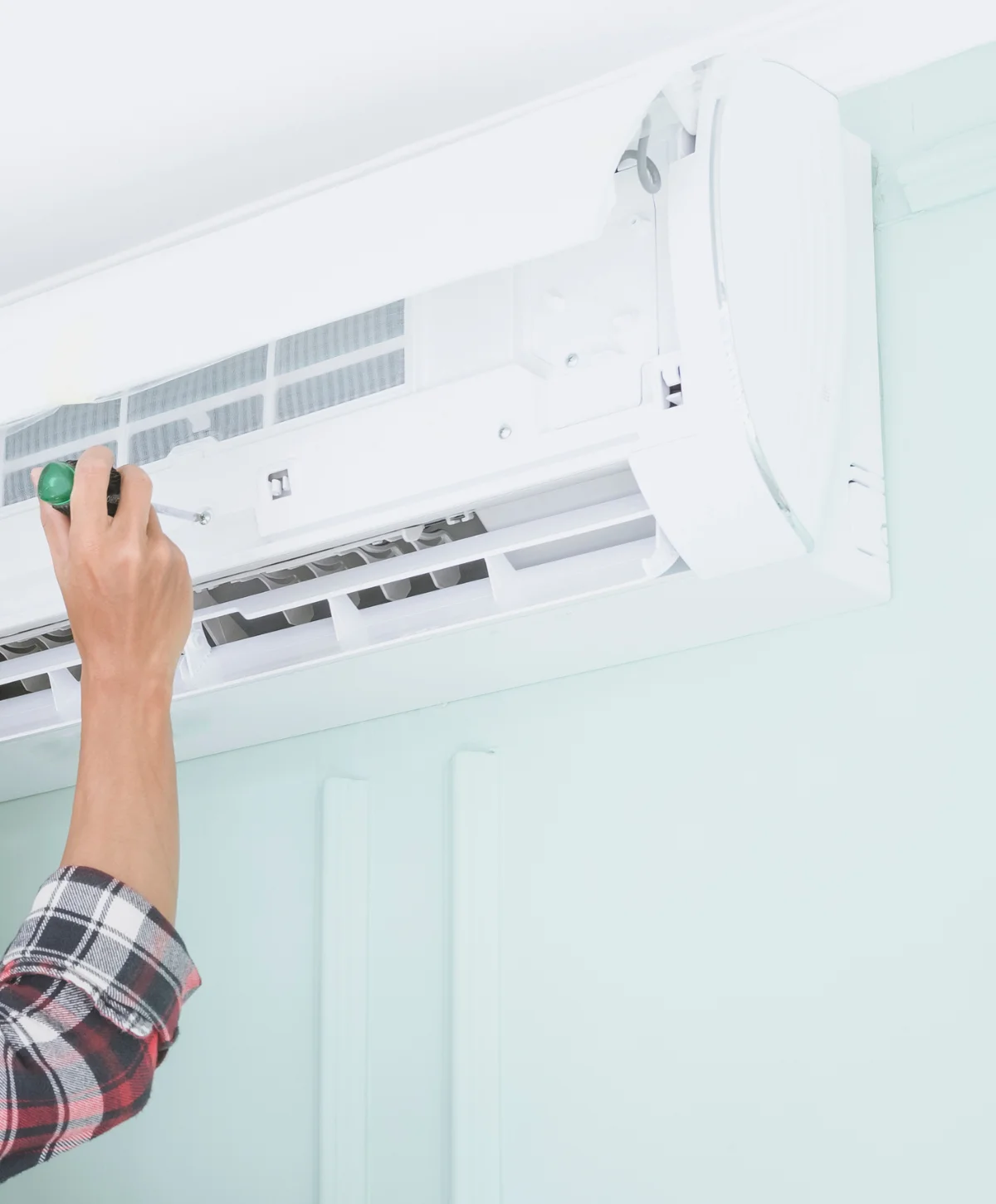 A person's hand in a plaid shirt uses a screwdriver to service an open white air conditioner indoor unit.