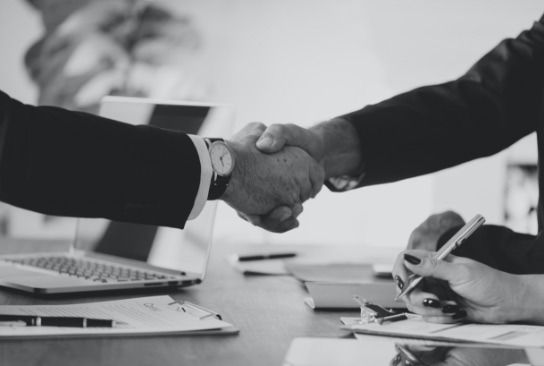 Black and white photograph of two people shaking hands across a desk holding a laptop and printed documents