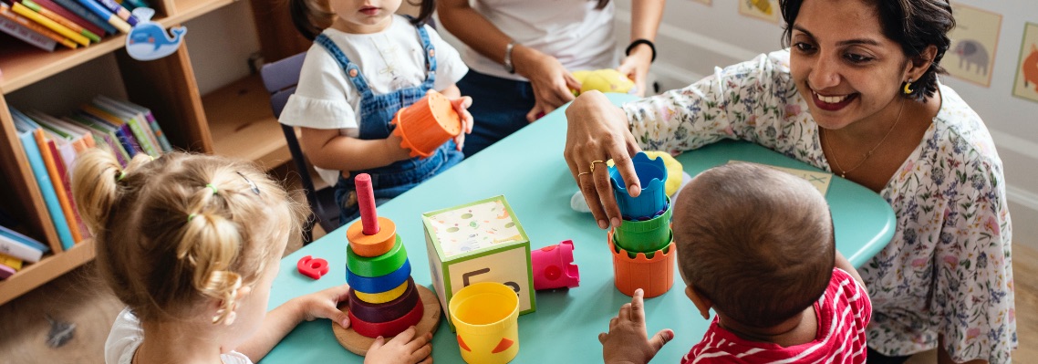 Preschool teacher guiding toddlers playing with colourful stacking toys in daycare classroom