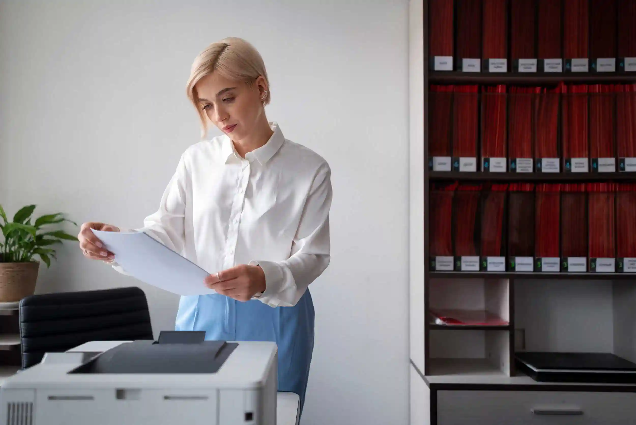 Woman standing at an office printer holding a printed document, shelves of lever-arch files behind her