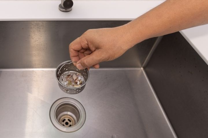 A hand holding a sink strainer filled with food debris to prevent a drain backup.