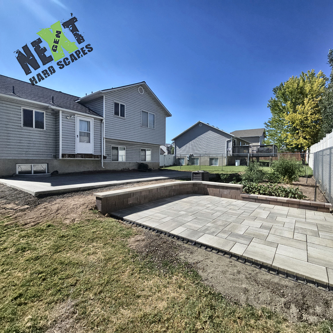 Step-down paver patio with newly laid interlocking pavers and a curved retaining wall bench bordering the vegetable garden.
