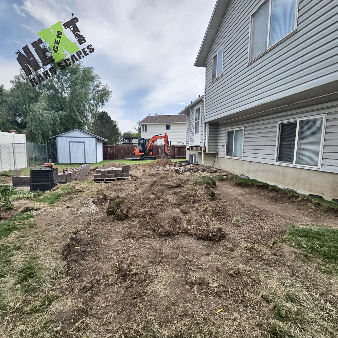 The backyard is cleared and prepped with excavation equipment and retaining wall blocks staged for installation near the vegetable garden.