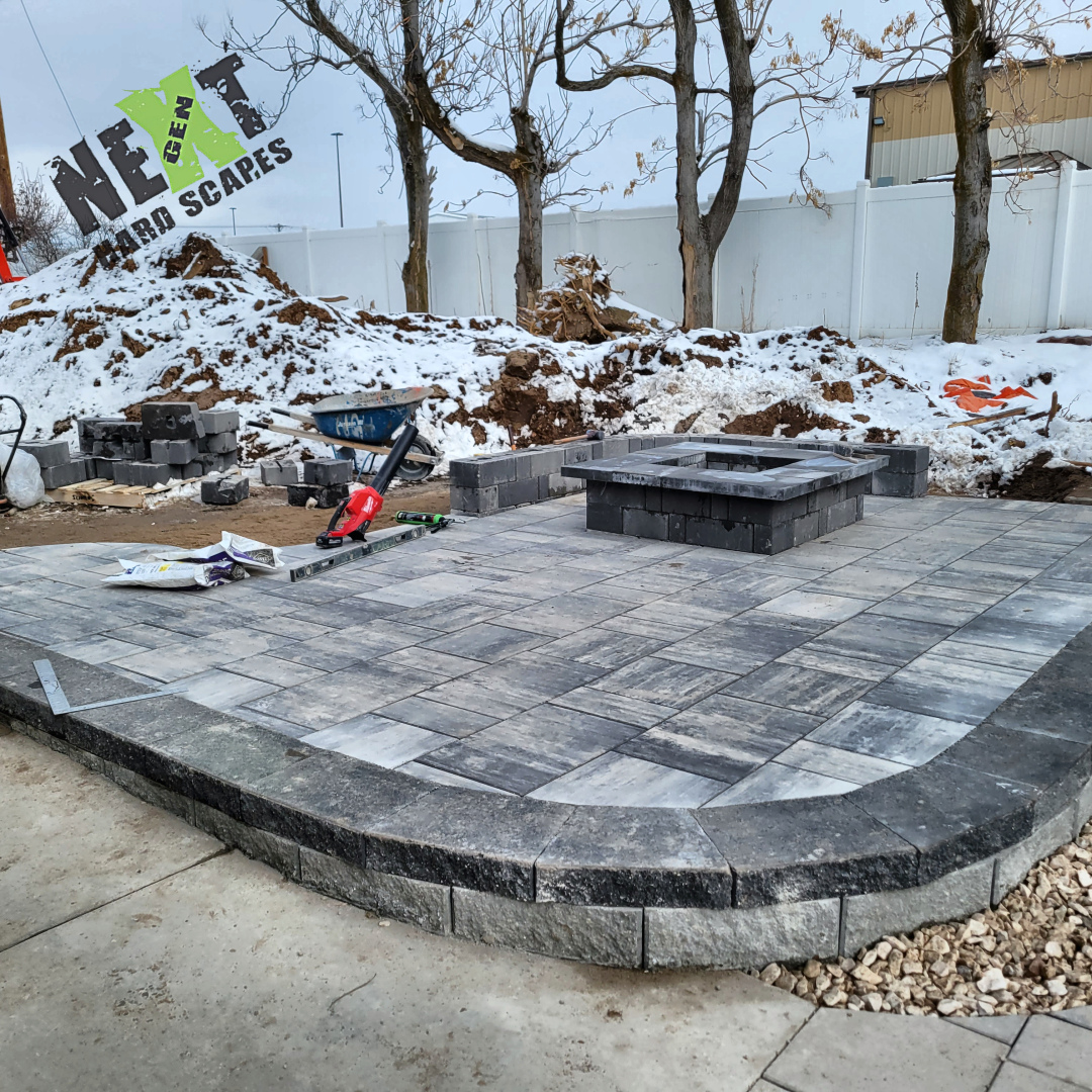 Paver patio mostly completed with defined edging and a raised fire pit base constructed in the center area. Paver patio mostly completed with defined edging and a raised fire pit base constructed in the center area.