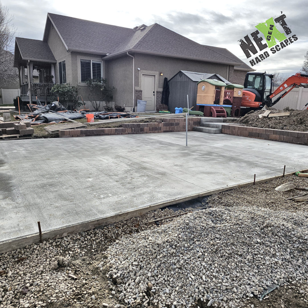 Finished concrete patio slab with a broomed surface and a vertical electrical conduit set in the center for a future pergola, bordered by the new retaining wall. Finished concrete patio slab with a broomed surface and a vertical electrical conduit set in the center for a future pergola, bordered by the new retaining wall.