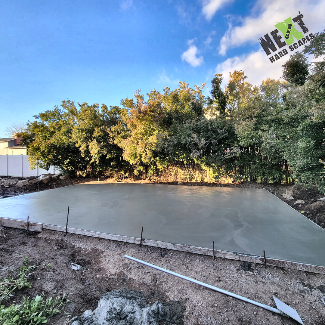 Backyard patio pad freshly poured and smoothed within straight timber forms, with stakes holding the perimeter in place.