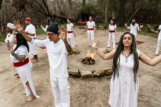 Group in white clothing standing in a circle around a small fire during an outdoor holistic ceremony.