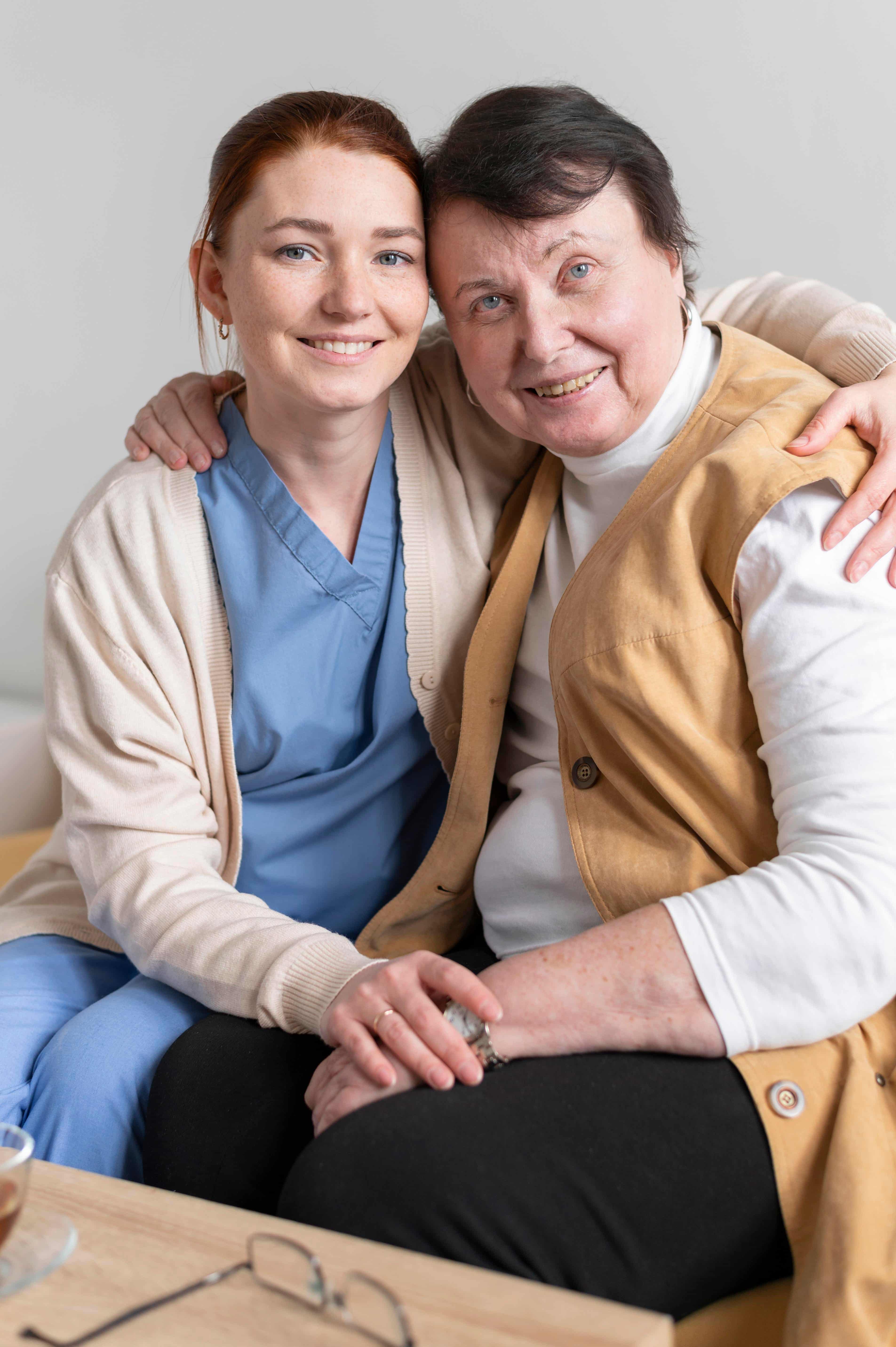 A smiling elderly man sitting in a chair at home, supported by a compassionate home care caregiver wearing a blue uniform and light cardigan in a bright, comfortable room.