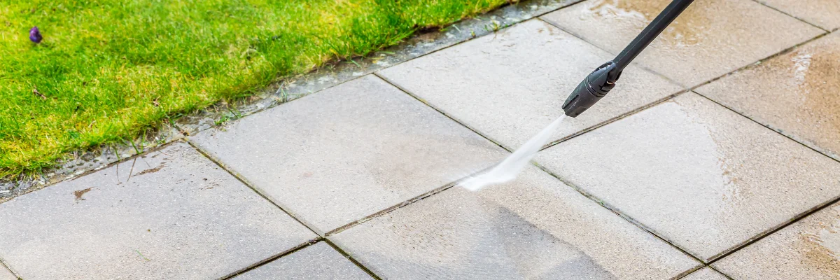 Pressure washer cleaning patio tiles beside green lawn.