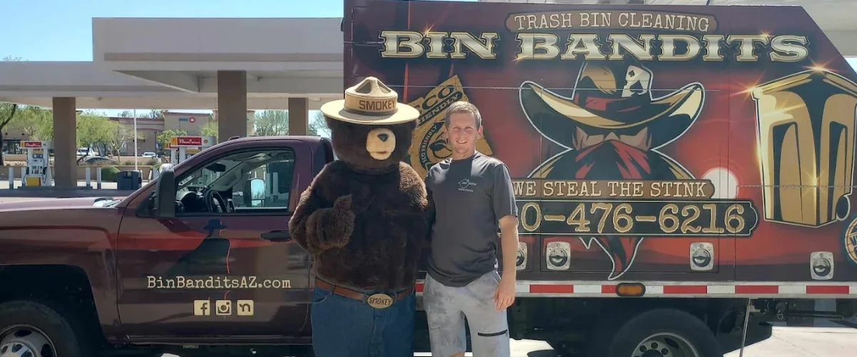 A man and Smokey Bear mascot standing next to a Bin Bandits trash bin cleaning truck with custom graphics.