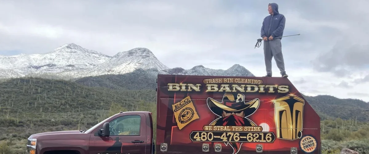 A man stands atop a red Bin Bandits trash bin cleaning truck, holding a hose, with snowy mountains in the background.