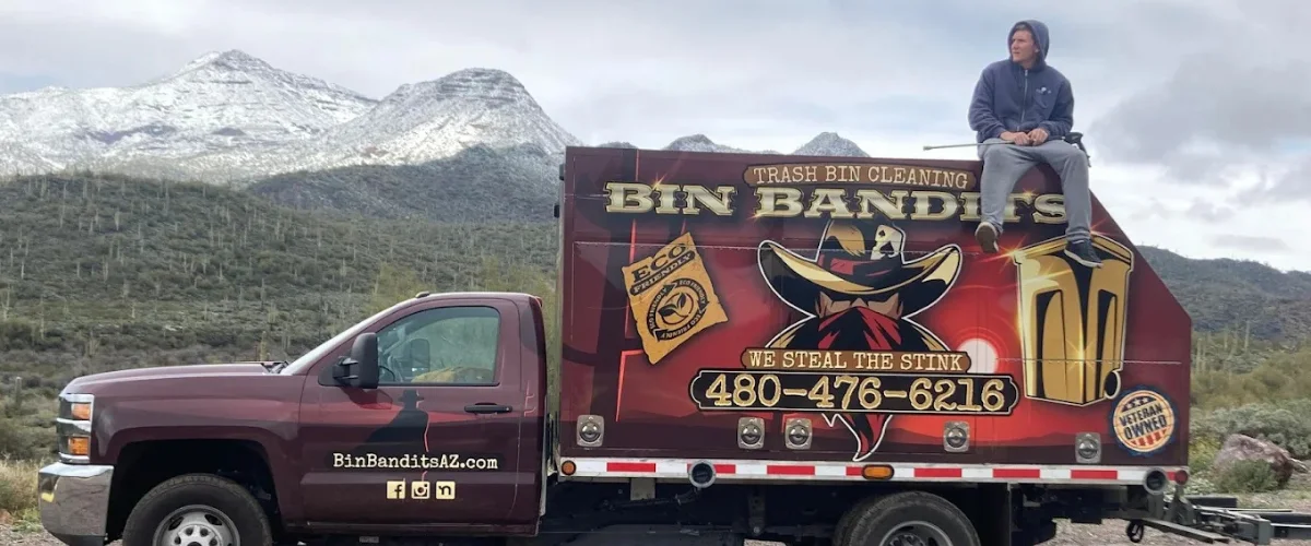 A maroon Bin Bandits trash bin cleaning truck with a man sitting on top, set against snowy mountains and desert landscape.