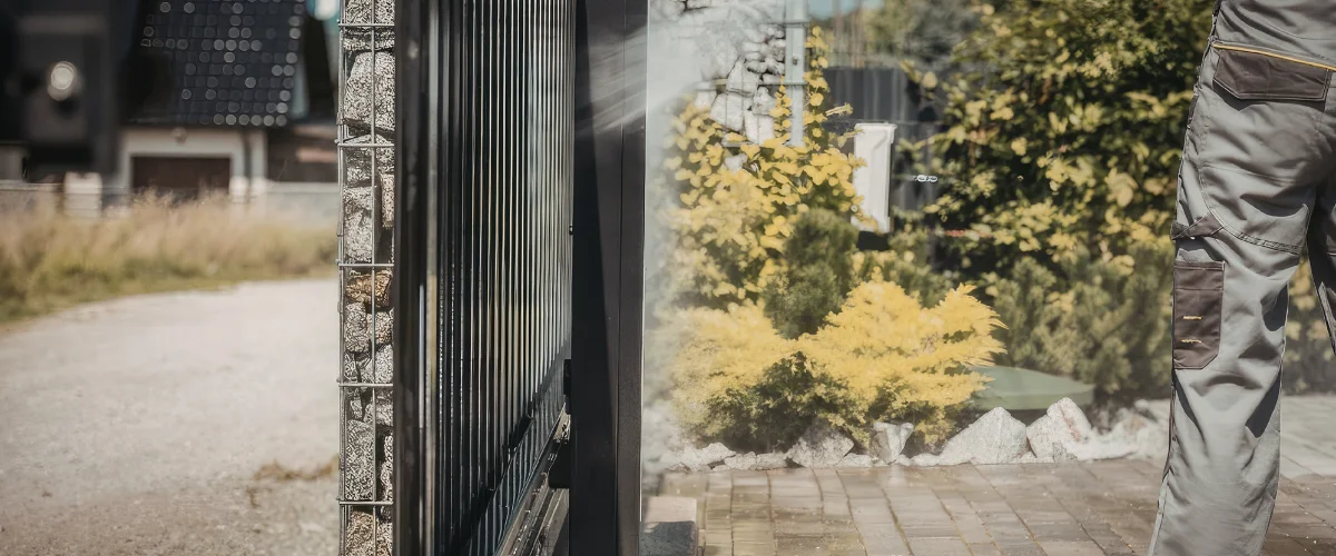 A close-up of a gabion wall and fence, with a person in work pants and foliage in the reflection on a sunny day.