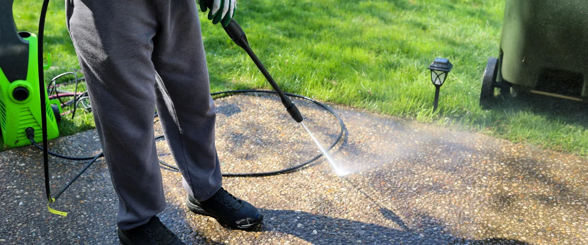A person in gray sweatpants and gloves power washing a concrete pebble aggregate patio or path. A person in gray sweatpants and gloves power washing a concrete pebble aggregate patio or path.
